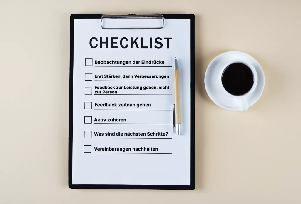 Eine Checkliste in deutscher Sprache auf einem Klemmbrett mit einem Stift und einer Tasse schwarzen Kaffees daneben, auf einer beigen Fläche platziert.