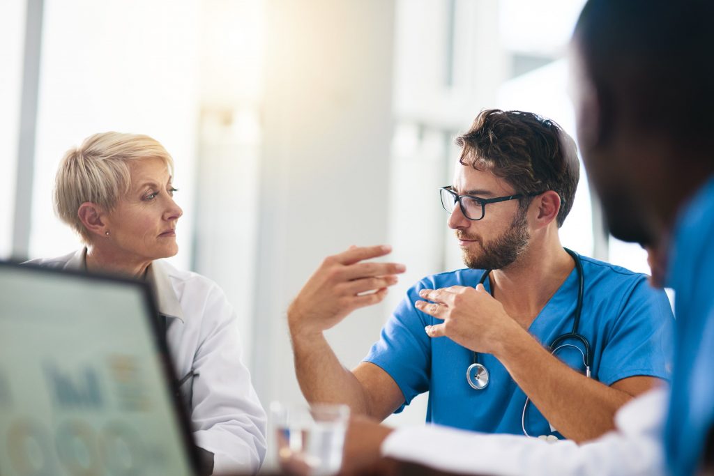 Eine Gruppe von Angehörigen der Gesundheitsberufe sitzt an einem Tisch und diskutiert; eine Person im blauen Kittel gestikuliert, während sie spricht.