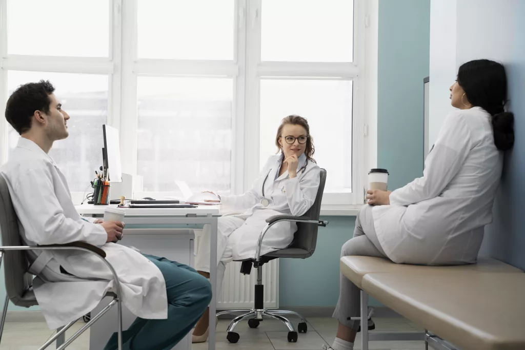Drei Mediziner in Laborkitteln sitzen in einem hellen Büro und unterhalten sich; einer sitzt an einem Schreibtisch mit einem Laptop, ein anderer auf einem Stuhl, und der dritte hält eine Kaffeetasse, während er auf einer Bank sitzt.
