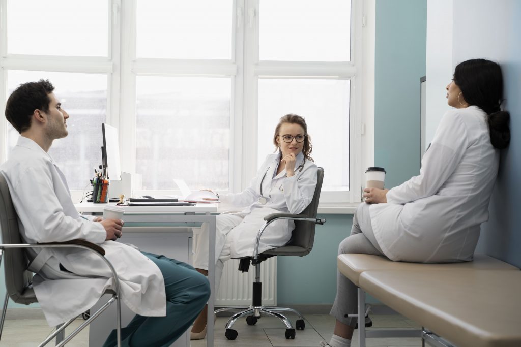 Drei Mediziner in Laborkitteln sitzen in einem hellen Büro und unterhalten sich; einer sitzt an einem Schreibtisch mit einem Laptop, ein anderer auf einem Stuhl, und der dritte hält eine Kaffeetasse, während er auf einer Bank sitzt.