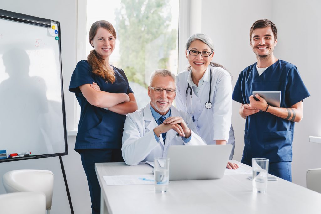 Vier Fachkräfte aus dem Gesundheitswesen, zwei Frauen und zwei Männer, posieren gemeinsam in einem hellen Büro mit Laptop, Whiteboard und Wassergläsern auf dem Tisch - ein Zeichen für starke Mitarbeiterbindung im Krankenhaus.