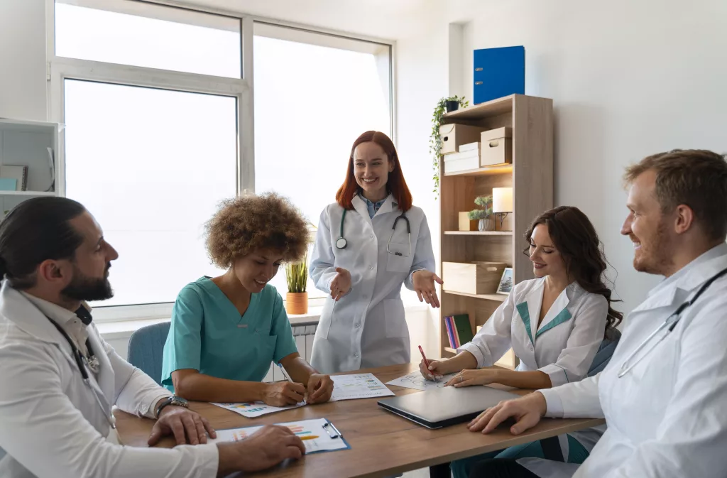 Fünf medizinische Fachkräfte in medizinischer Kleidung sitzen und stehen um einen Tisch in einem Büro, besprechen Dokumente und lächeln während einer Teambesprechung.