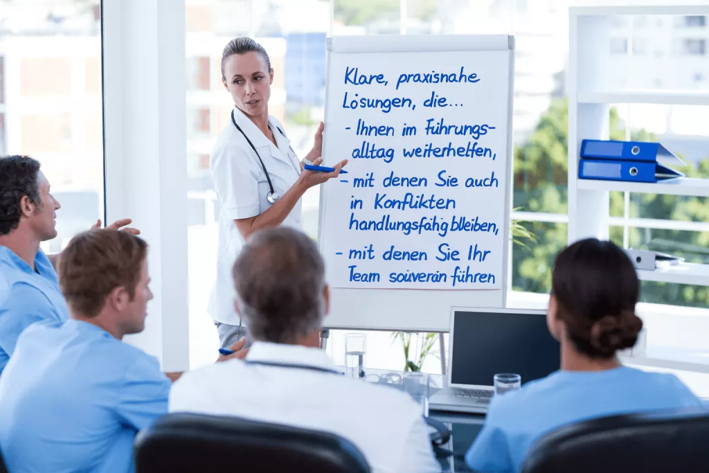 Eine Frau in medizinischer Kleidung hält einen Vortrag vor Kollegen und zeigt auf ein Flipchart mit einem deutschen Text über Führungslösungen im Gesundheitswesen. Vier sitzende Personen hören aufmerksam zu.